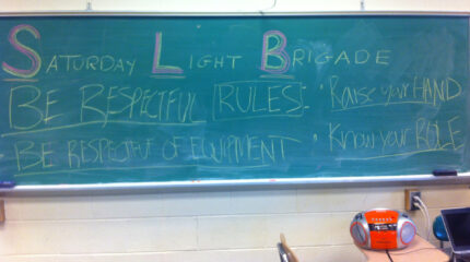 Yellow chalk on a green chalkboard detailing basic classroom etiquette for the Saturday Light Brigade