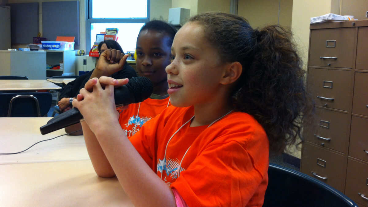 A youth looking at another youth holding a microphone with two hands, both sitting at classroom desks