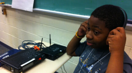 A youth wearing a pair of headphones sitting behind a tape recorder on a classroom desk