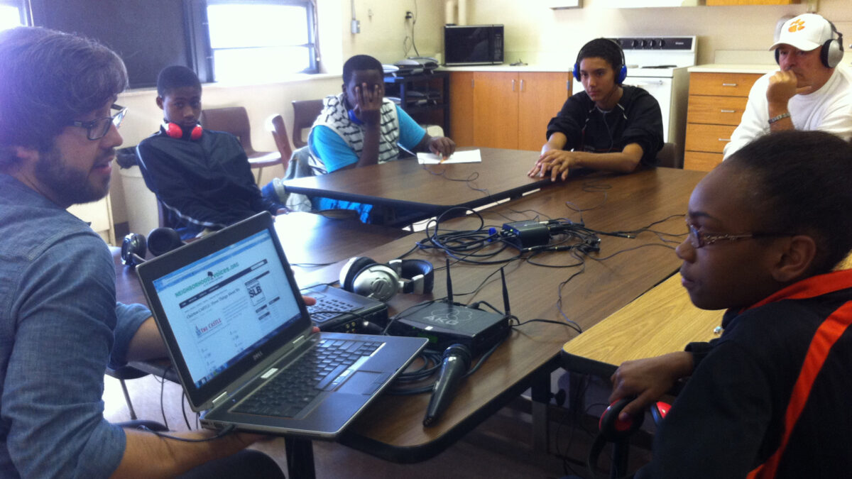 An adult showing a youth a laptop computer as three youth and an adult wearing headphones sitting around a long brown table watch in a classroom