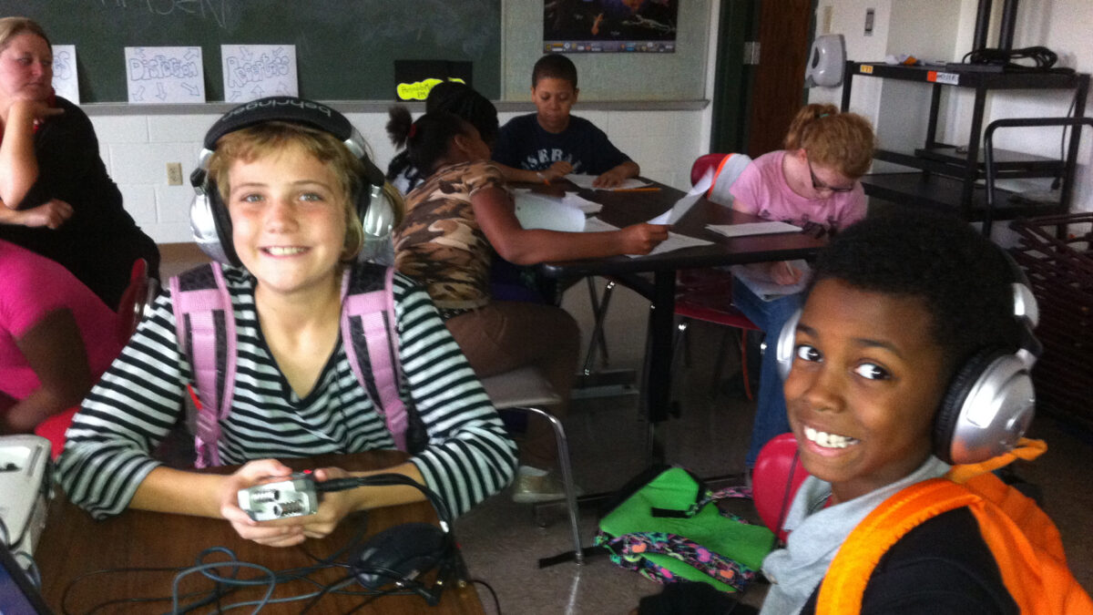 Two youth sitting at a classroom table wearing headphones smiling at the camera, one holding a portable recorder, and four youth sitting at a classroom table looking at pieces of paper in the background