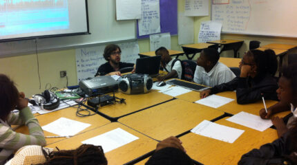 An adult sitting behind a laptop computer showing a class of youth audio editing software in a classroom