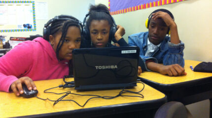 Three teens wearing headphones looking at a laptop computer on a classroom desk