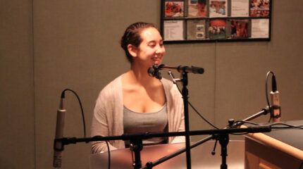 A teen smiling while sitting behind three microphones in a radio studio