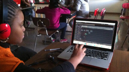 A youth wearing headphones interacting with audio editing software on a laptop on a classroom desk