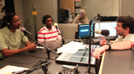 An adult and a teen wearing headphones speaking into microphones being interviewed by an adult sitting behind a microphone and a mixer in a radio studio