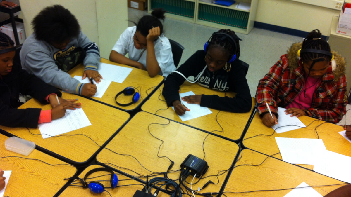 Five youth, two wearing headphones, writing on pieces of paper on classroom desks pushed together
