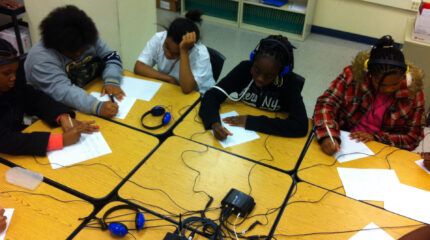 Five youth, two wearing headphones, writing on pieces of paper on classroom desks pushed together