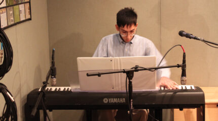 A teen playing a piano keyboard sitting behind a binder and three microphones in a radio studio