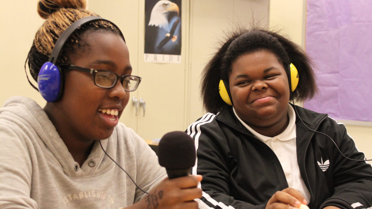 Two teens wearing headphones speaking into a microphone in a classroom.