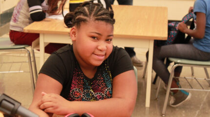 A youth sitting behind a microphone and a pair of headphones at a classroom desk