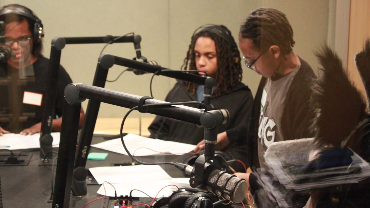 Four youth, three wearing headphones, sitting behind pieces of paper and microphones in a radio studio