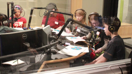 Five youth wearing headphones sitting behind pieces of paper and microphones in a radio studio