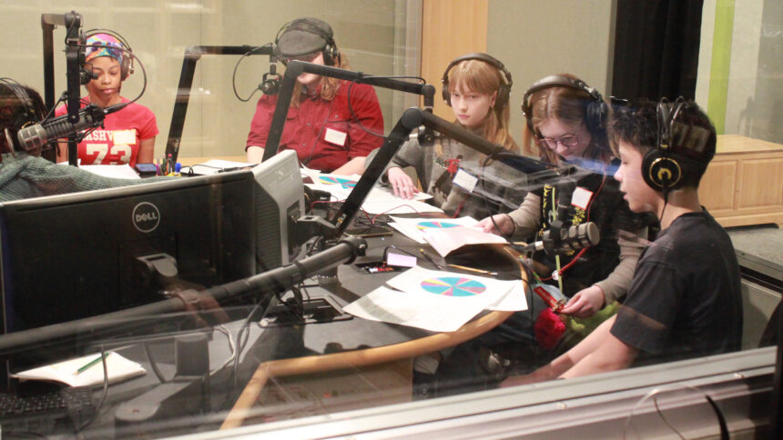 Five youth wearing headphones sitting behind pieces of paper and microphones in a radio studio