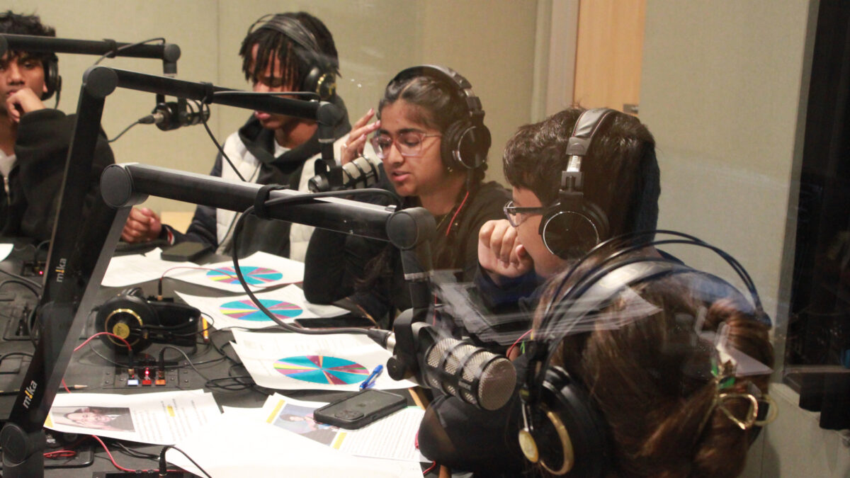 Five youth wearing headphones sitting behind pieces of paper and speaking into microphones in a radio studio