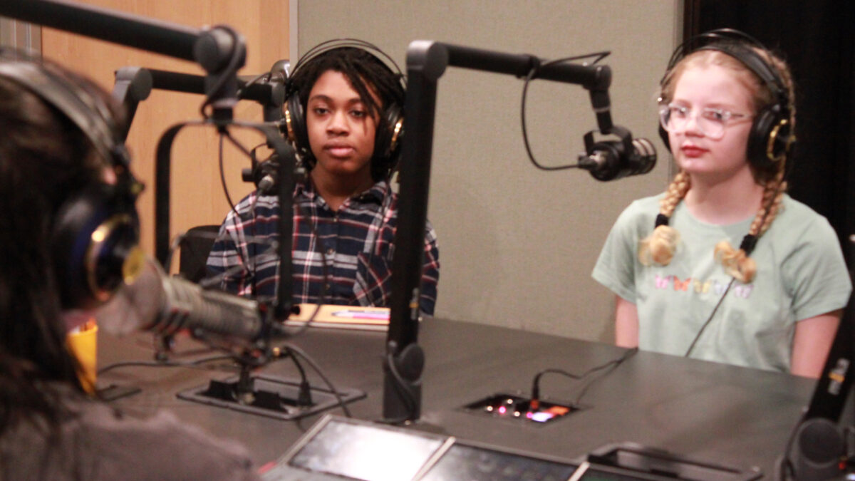 Two youth wearing headphones sitting behind microphones and an adult wearing headphones sitting behind a microphone and an audio mixer in a radio studio