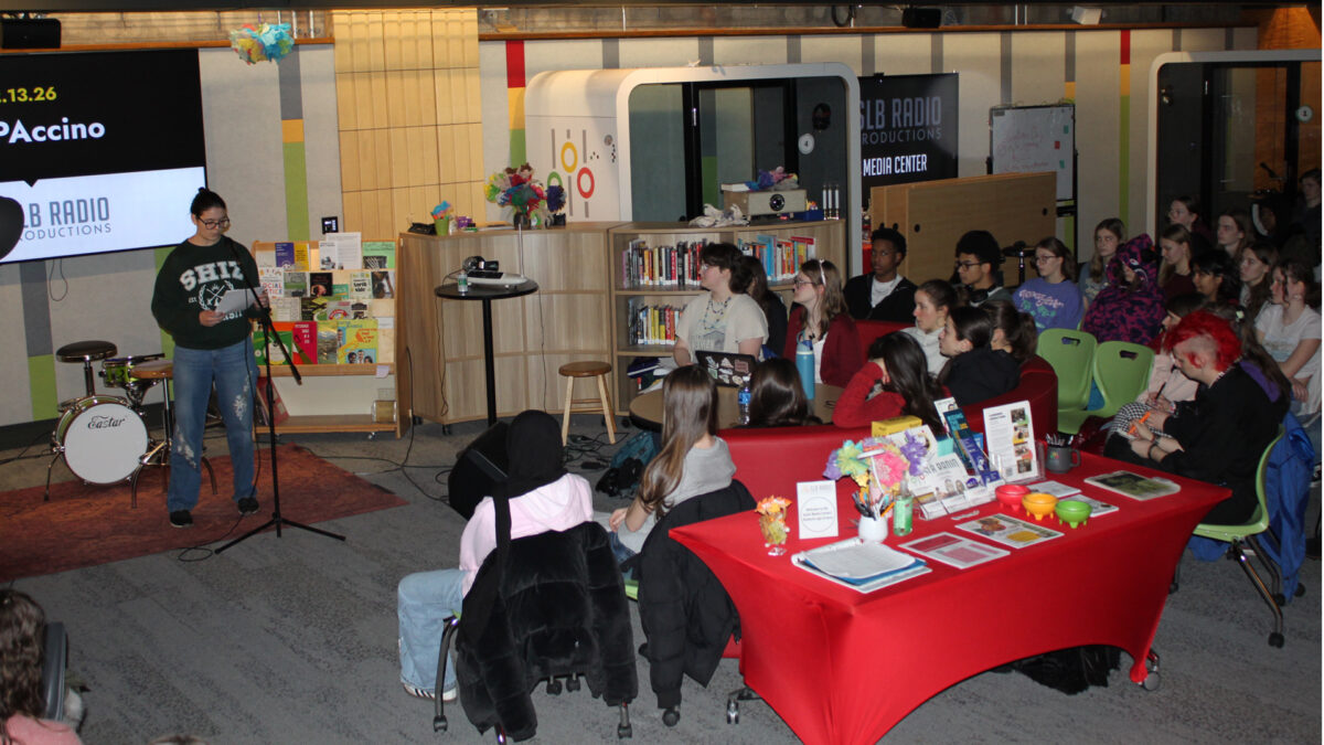 Twenty two teens listening to another teen reading a piece of paper into a microphone