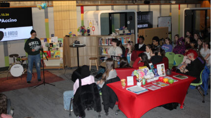 Twenty two teens listening to another teen reading a piece of paper into a microphone