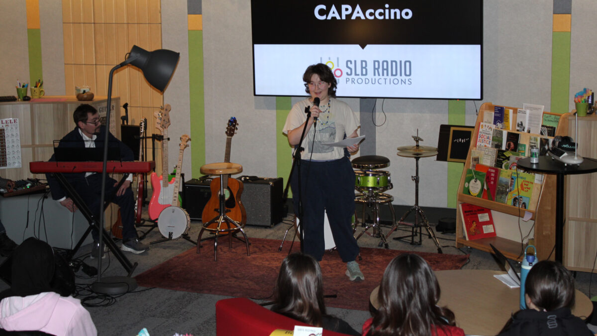 Four teens and an adult listening to a teen reading from a piece of paper into a microphone