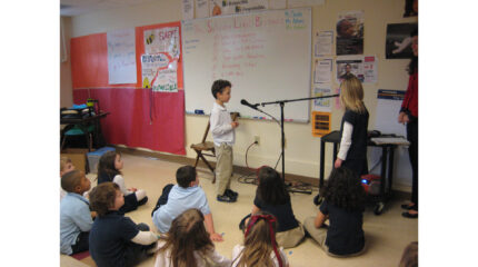 A youth standing and speaking into a microphone in front of a whiteboard as ten other youth, one standing and the rest sitting on the floor, listen in a classroom