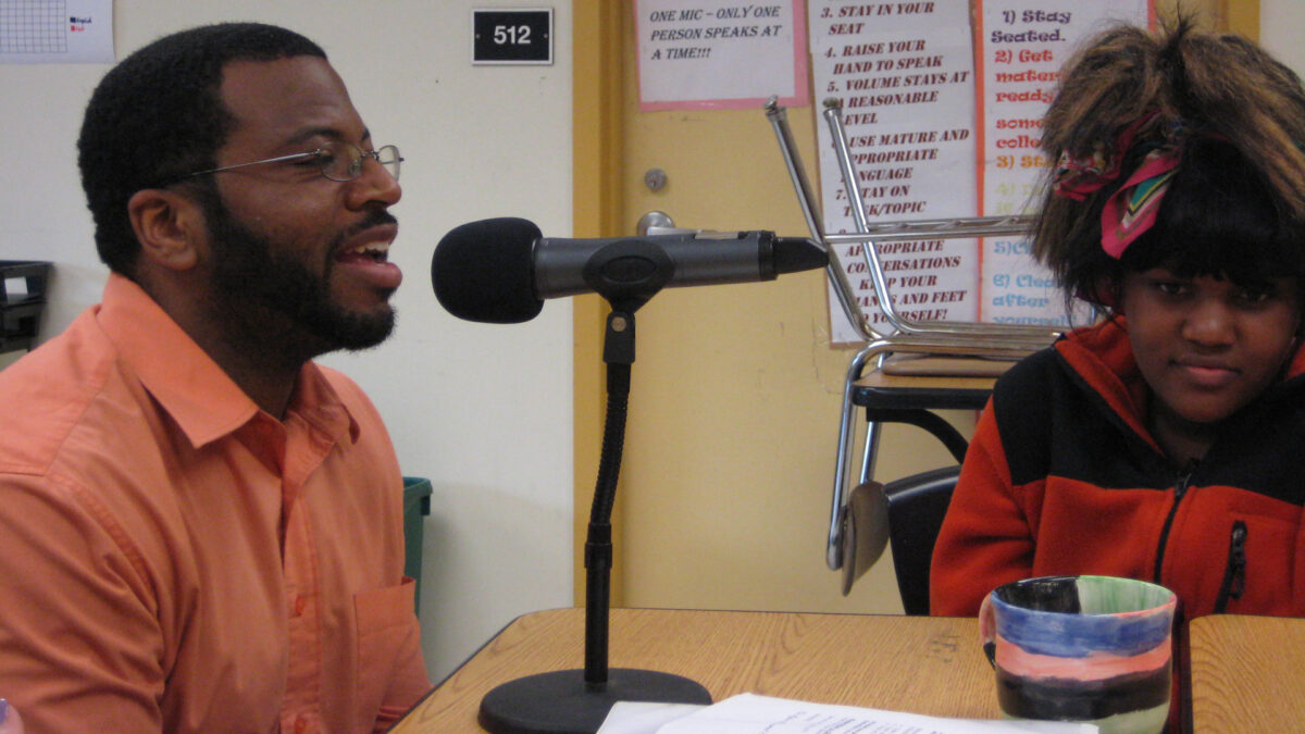 A teen listening to an adult speaking into a microphone, both sitting at a classroom table