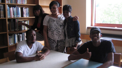 Five teens posing for the camera in a library, one smiling and flashing a thumbs up