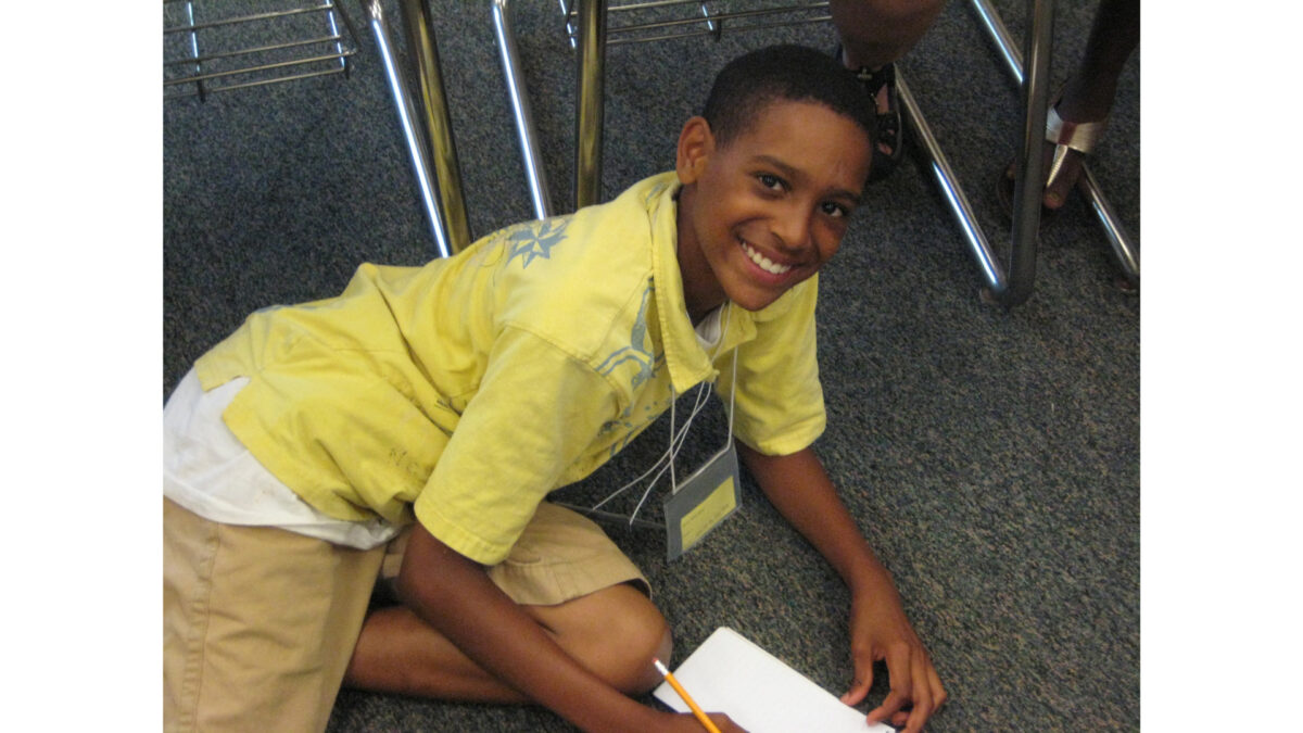 A youth sitting on the floor smiling at the camera and writing on a piece of paper