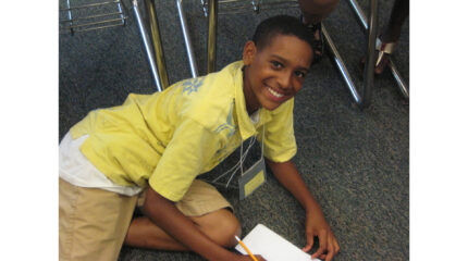 A youth sitting on the floor smiling at the camera and writing on a piece of paper