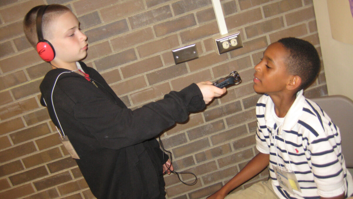 A youth wearing headphones holding a portable recorder interviewing another youth sitting