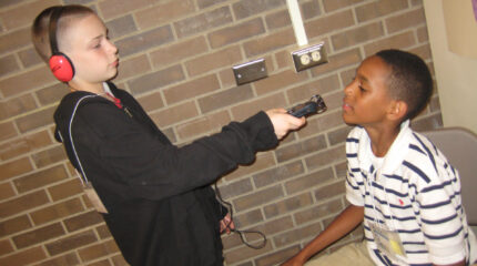 A youth wearing headphones holding a portable recorder interviewing another youth sitting