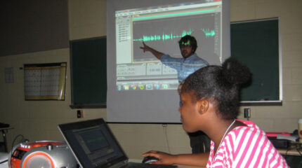 An adult pointing at a projected screen of audio editing software and a youth looking at a laptop computer in a classroom.