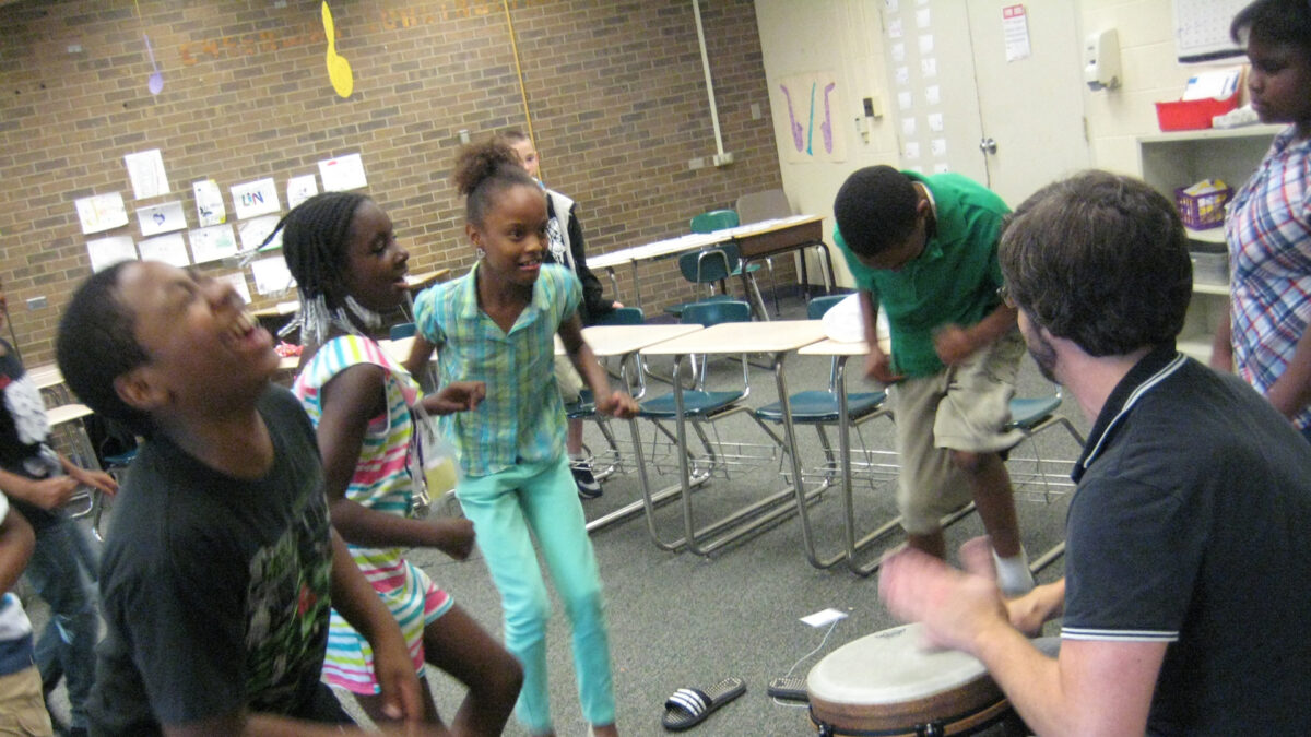 An adult playing a drum while five youth dance in a classroom