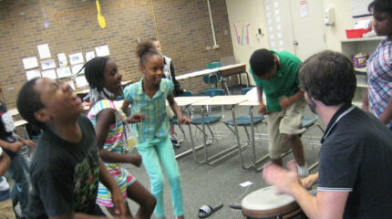 An adult playing a drum while five youth dance in a classroom