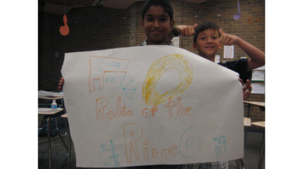 Two youth holding up a large piece of paper with drawings of buildings, swords, bows and arrows, and a large gold ring, and the text "Radio of the Rings."