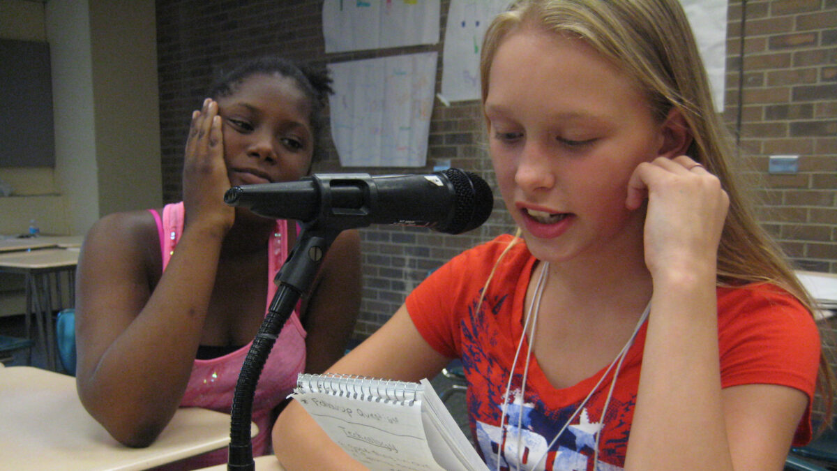 A youth looking at another youth holding a notepad and speaking into a microphone, both sitting at classroom desks