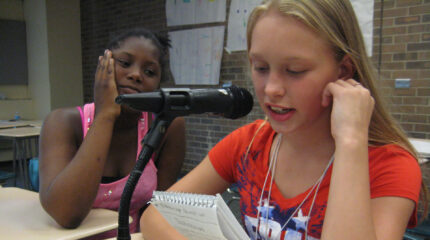 A youth looking at another youth holding a notepad and speaking into a microphone, both sitting at classroom desks