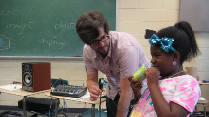 An adult holding a wire looking at a youth holding a light green plastic cup in a classroom