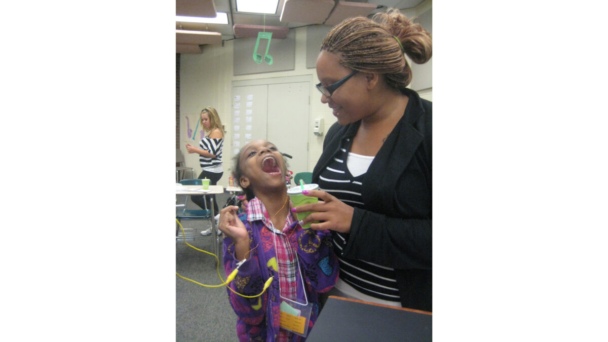 A youth with their mouth wide open and an adult holding a green plastic cup looking at the youth in a classroom