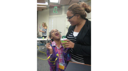 A youth with their mouth wide open and an adult holding a green plastic cup looking at the youth in a classroom