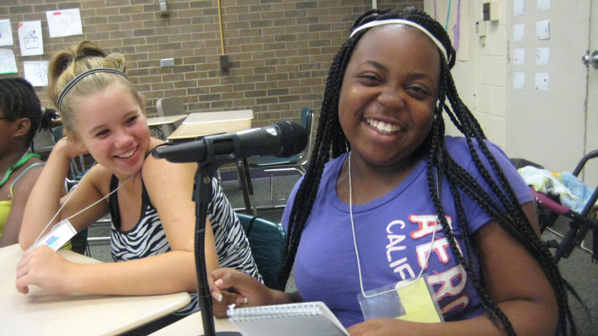 Two youth smiling at the camera while sitting at classroom desks, one holding a notepad and sitting behind a microphone