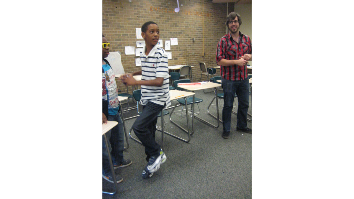 An adult wearing glasses and a youth wearing sunglasses watching another youth assume a pitching stance in a classroom