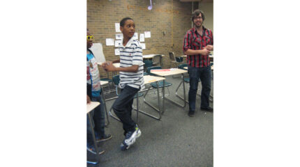 An adult wearing glasses and a youth wearing sunglasses watching another youth assume a pitching stance in a classroom