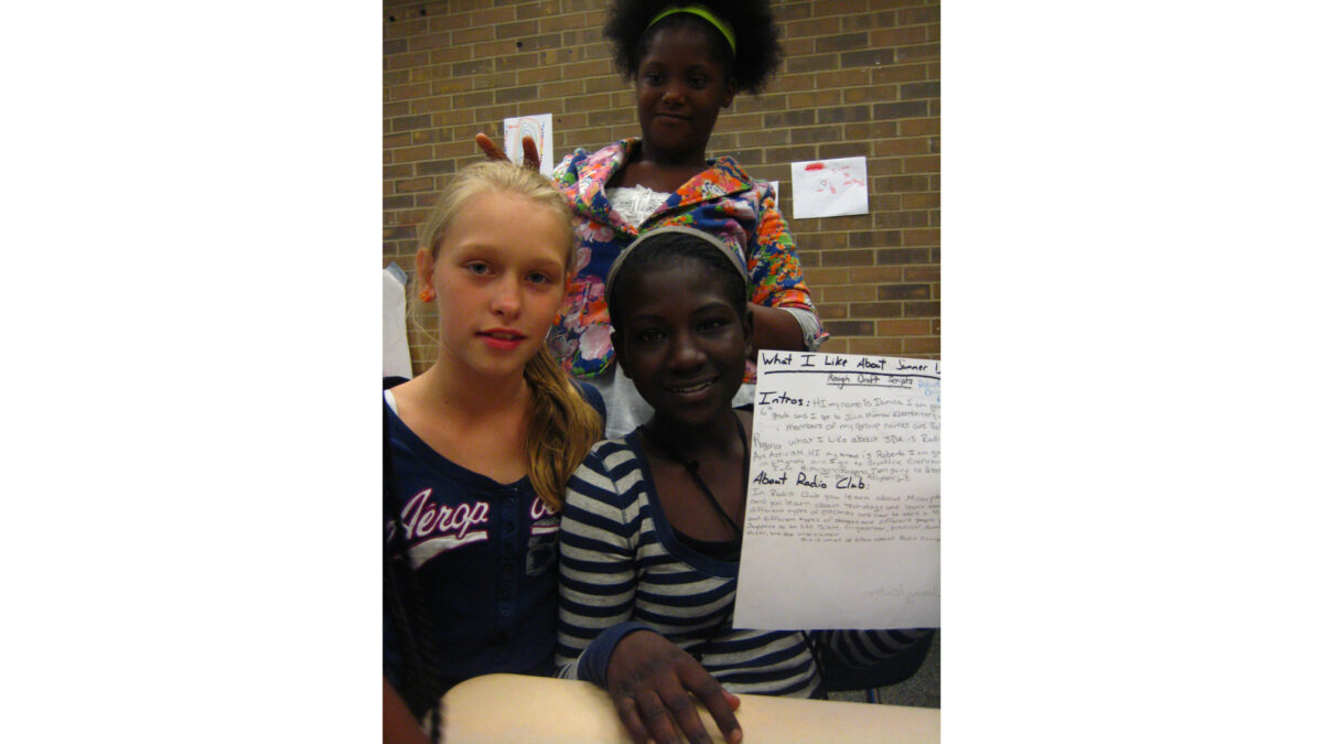 Three youth sitting and posing behind a classroom desk, one youth holding up a piece of paper with writing on it