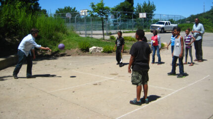 An adult tossing a kickball at six youth in a game of four square