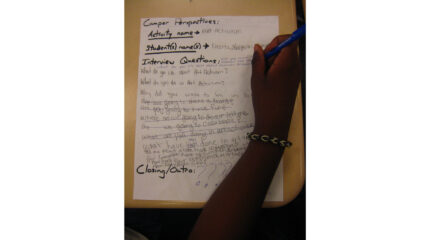 An individual's hand writing on a piece of paper on a classroom desk