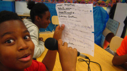 A youth making a shocked face at the camera while holding a piece of paper and a microphone and sitting at a classroom table