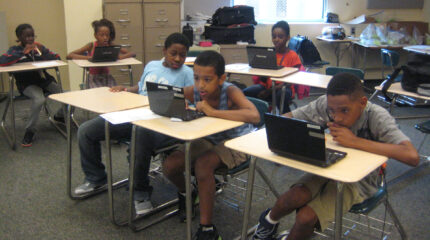 Six youth looking at laptop computers while sitting at classroom desks