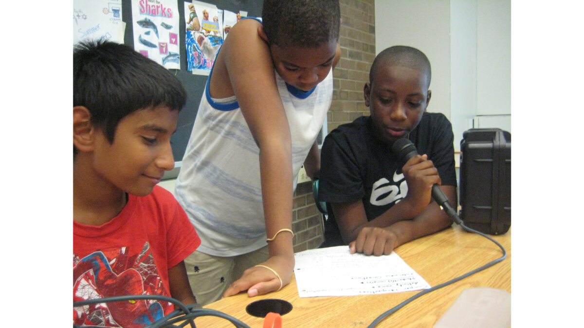 Three youth looking down at a piece of paper on a classroom table, one holding and speaking into a microphone