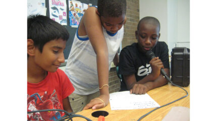 Three youth looking down at a piece of paper on a classroom table, one holding and speaking into a microphone
