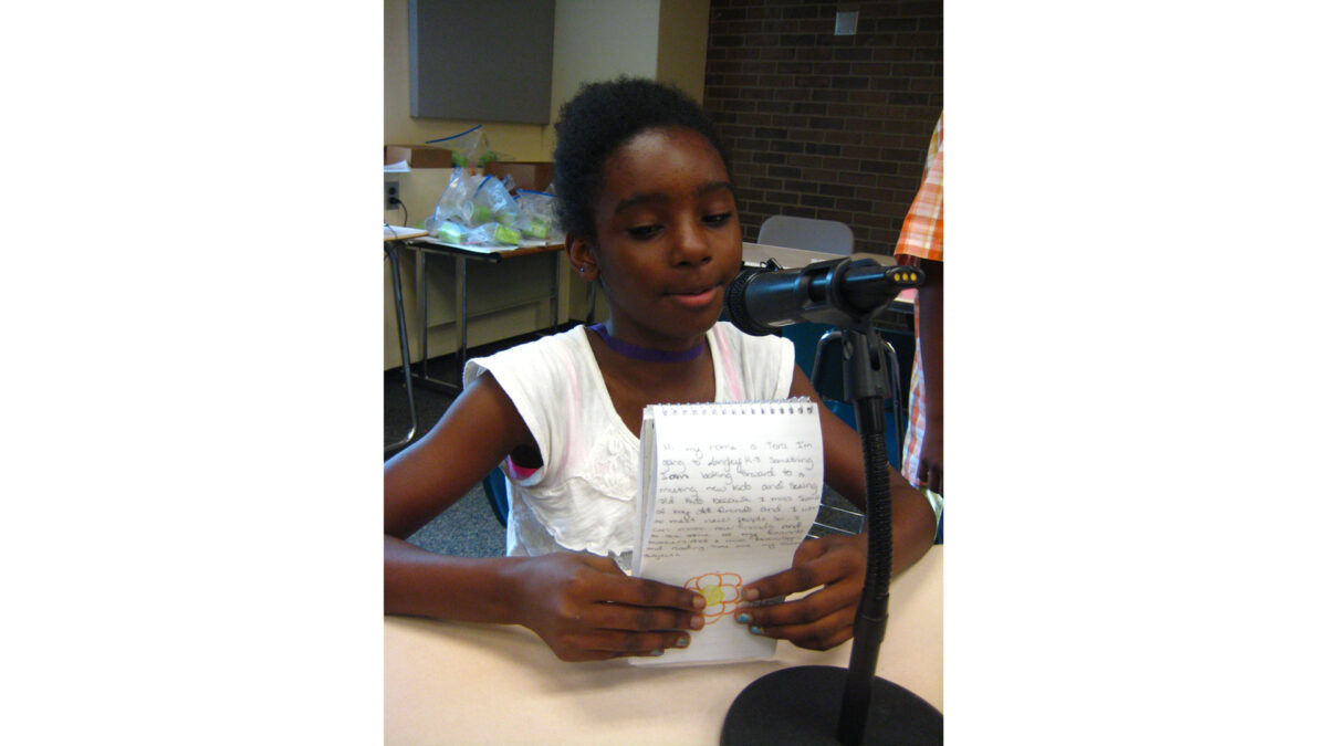 A youth looking at a notepad and speaking into a microphone sitting at a classroom desk